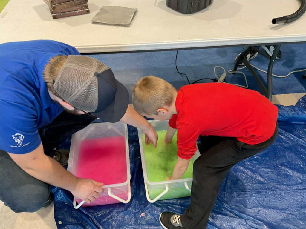 A Domtar mill employee helps a young student mix colorful pulp in large plastic tubs during a hands-on papermaking activity.