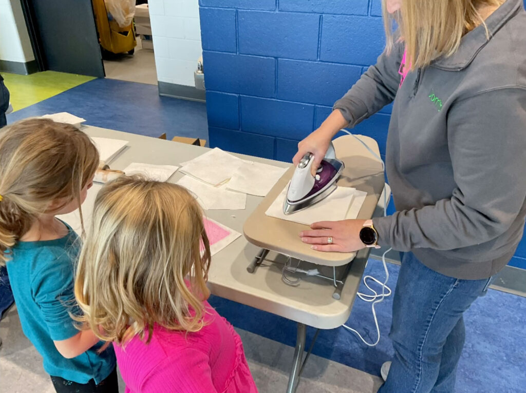 A Domtar employee uses an iron to dry handmade sheets of paper while two first graders watch during the classroom papermaking demonstration.